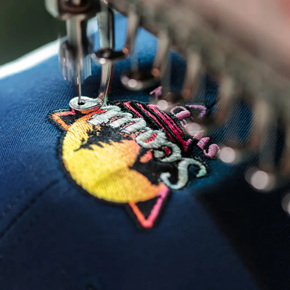 The industrial embroidery machine is stitching a vibrant, circular logo onto a blue cap